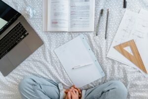 Flat lay of a study area with an open laptop, books, notes, and stationery on a bed.